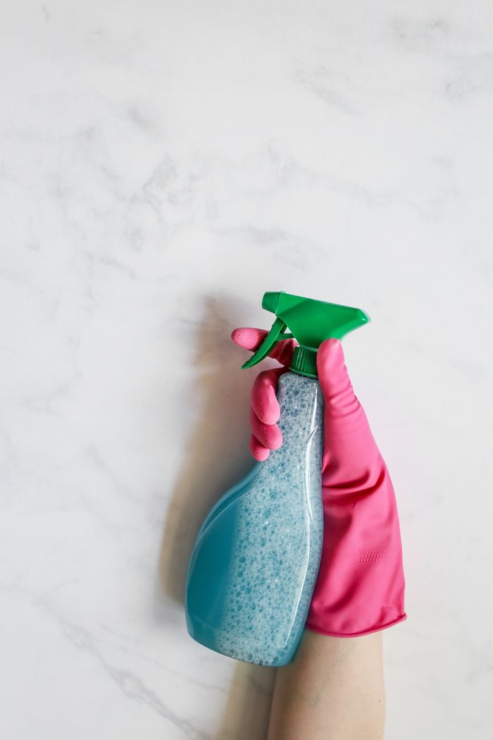 mobile-01 Close-up image of a hand in a pink glove holding a spray bottle against a white background.
